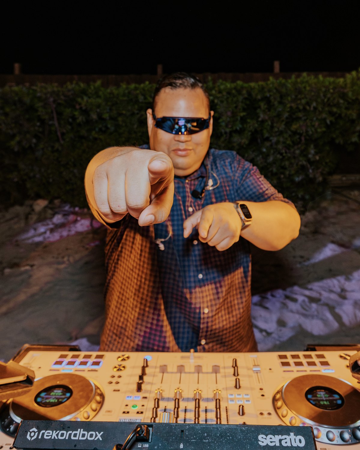 DJ Ruuben behind the decks at an outdoor celebration, pointing toward the camera.